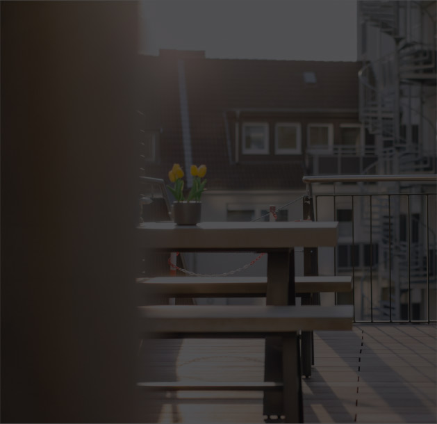 Holztisch mit gelben Tulpen auf einer Dachterrasse bei Abendsonne, umgeben von Wohnhäusern und Metallgeländer